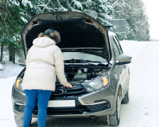 A person in a puffy white coat stands in front of the open hood of a car on a snowy road.